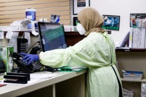 Pharmacy worker reaching for telephone while workinig on a laptop.